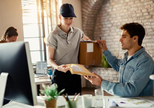 Smiling female courier delivering package to a businessman while he is working in the office.
